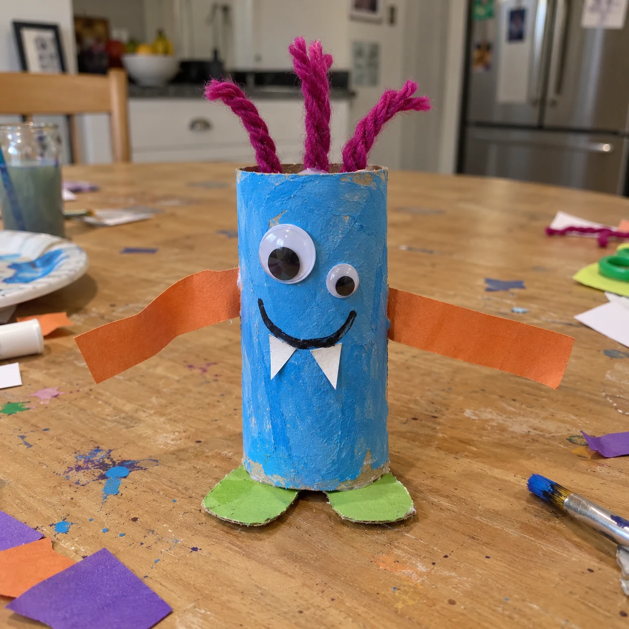 A handmade blue cardboard monster with yarn hair on a messy craft table